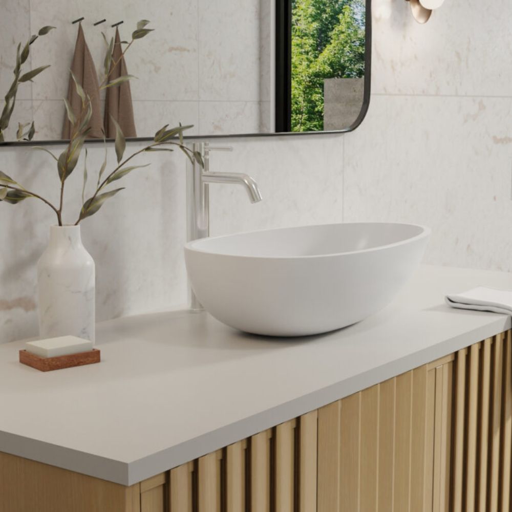 A modern bathroom vanity setup featuring a white oval vessel sink on a white countertop with a brushed metal faucet, set against a light stone tile wall with a mirror and decorative vase.