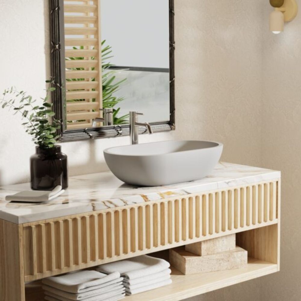 A modern floating vanity with a slatted wood front and marble countertop, holding a white oval vessel sink and chrome faucet. A dark-framed mirror, folded white towels, and a small plant in a dark vase are visible against a warm beige wall.