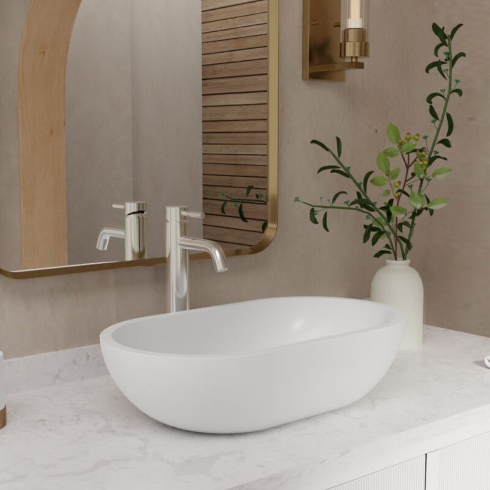 A white oval vessel sink basin set into a white marble bathroom vanity countertop, with a chrome faucet, a wall mirror, a gold-framed mirror, and a white vase with green branches on the counter.