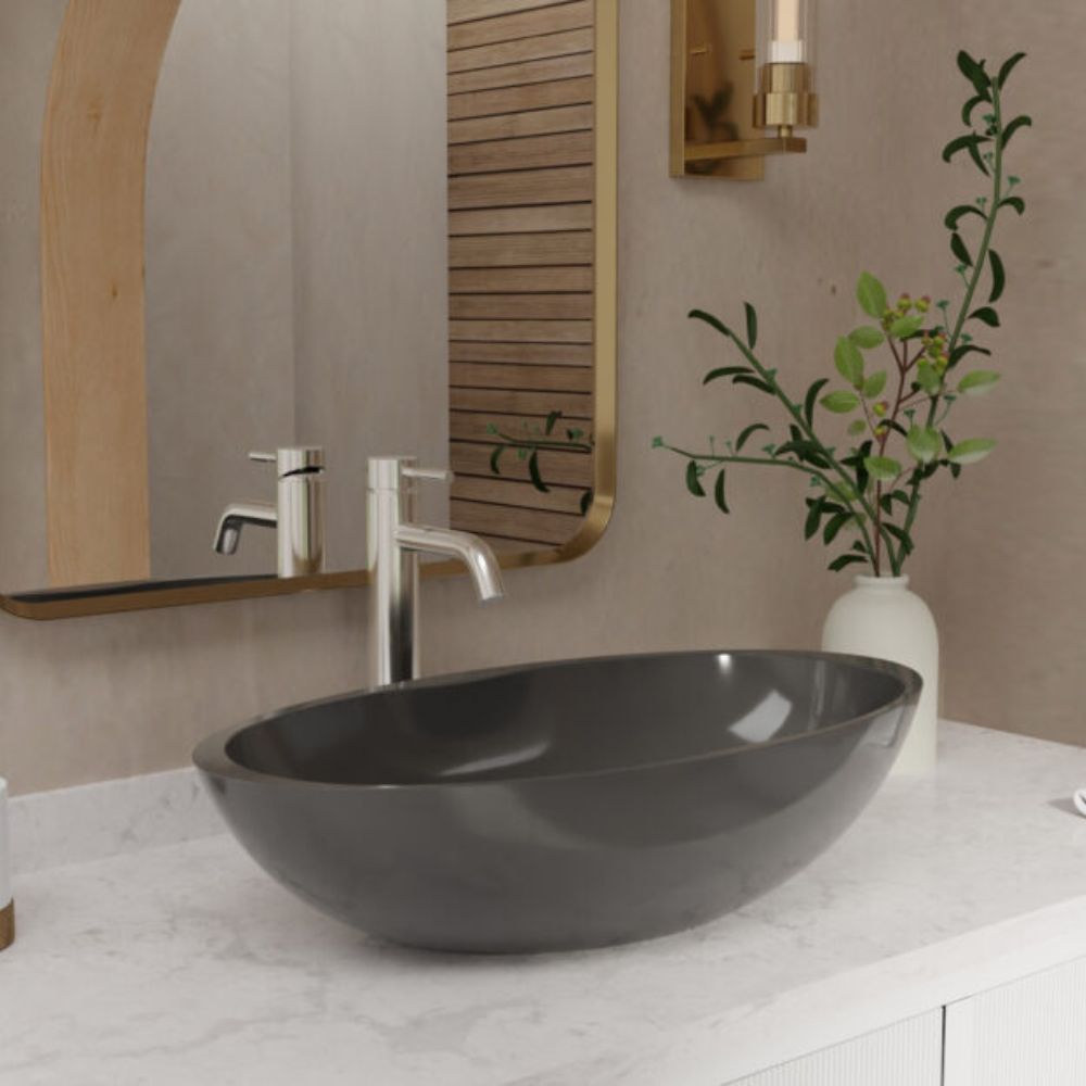 A dark charcoal gray vessel sink basin set into a white marble bathroom countertop with a modern chrome faucet, a gold-framed mirror, and a white vase with green leaves on a beige wall.