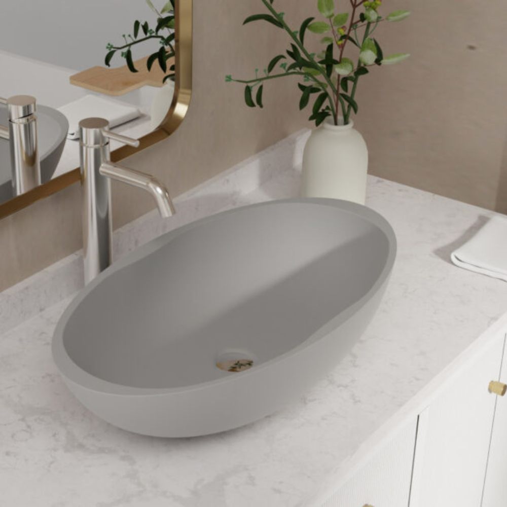 A modern oval gray vessel sink basin set into a white marble bathroom vanity countertop with a chrome faucet, mirror, and a white vase with greenery.
