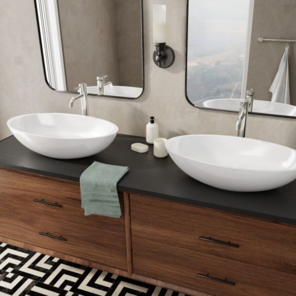 A modern bathroom vanity setup with two white oval vessel sinks on a dark countertop above a wood cabinet, with mirrors, faucets, soap dispensers, and a folded green towel visible.