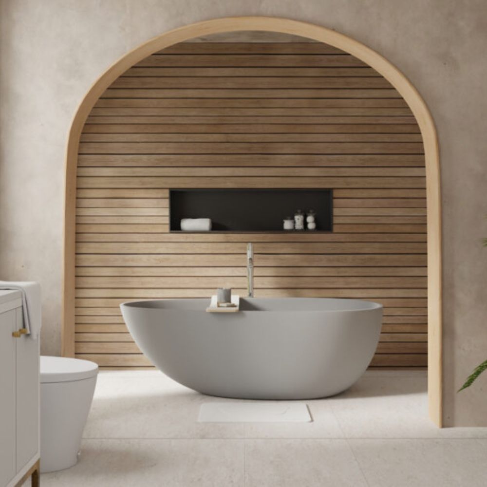 A modern freestanding oval bathtub in a minimalist bathroom setting, framed by a large arched wooden wall feature with a built-in niche and small toiletries.