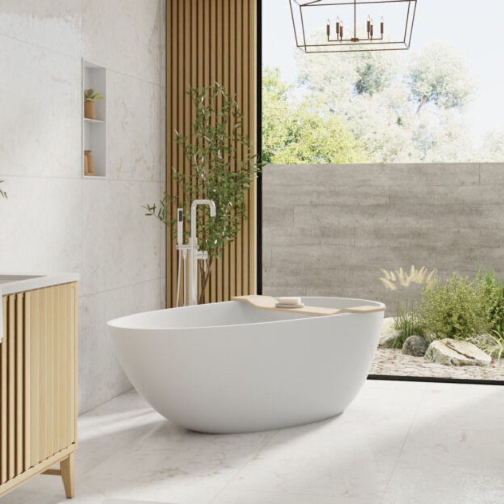 A modern freestanding oval bathtub in matte white, placed in a minimalist bathroom with light wood slat cabinetry, a vertical wood slat wall panel, and a large window looking out to greenery.