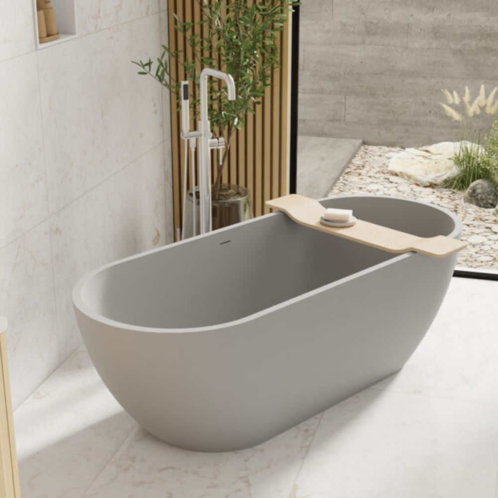 A modern freestanding oval bathtub in matte gray, shown in a styled bathroom setting with a matching chrome faucet, a wooden shelf insert holding a soap dish, and a spa-like outdoor-looking zen corner in the background.