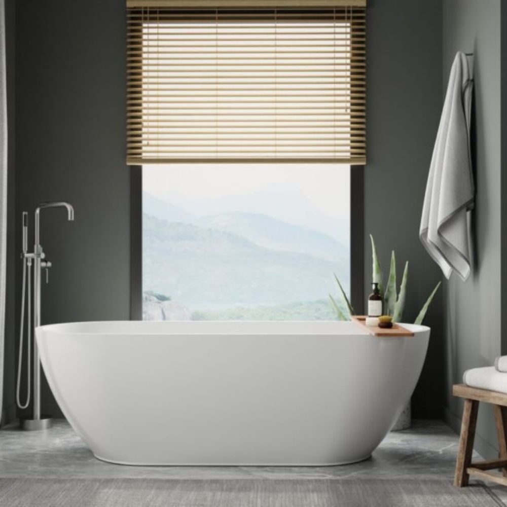 A modern freestanding oval bathtub in a dark gray bathroom, positioned in front of a window with wooden blinds and an outdoor mountain view. A chrome floor-mounted shower is on the left, a towel hangs on the right wall, and a small wooden stool with a plant sits beside the tub.