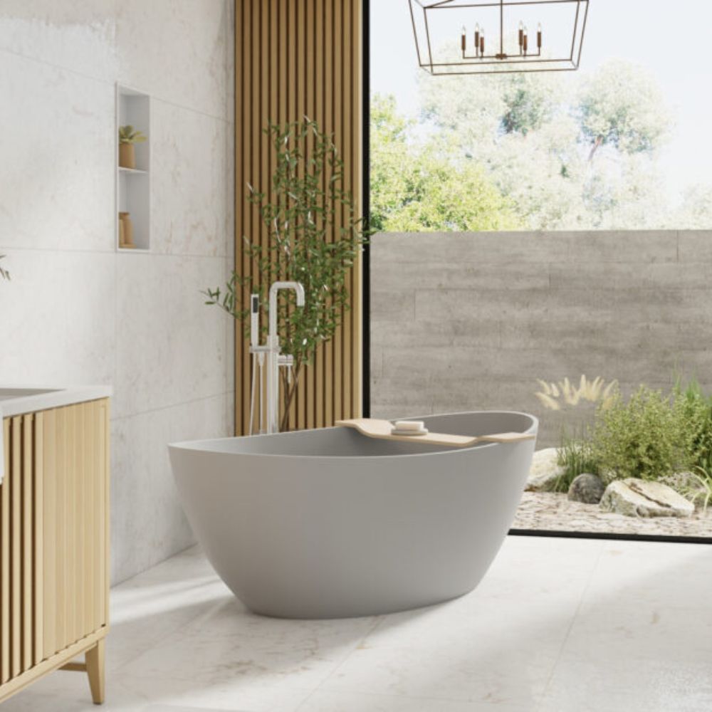 A modern freestanding oval bathtub in a light gray color, set in a contemporary bathroom with a wooden slat vanity, tiled walls, and a large window looking out to a garden.