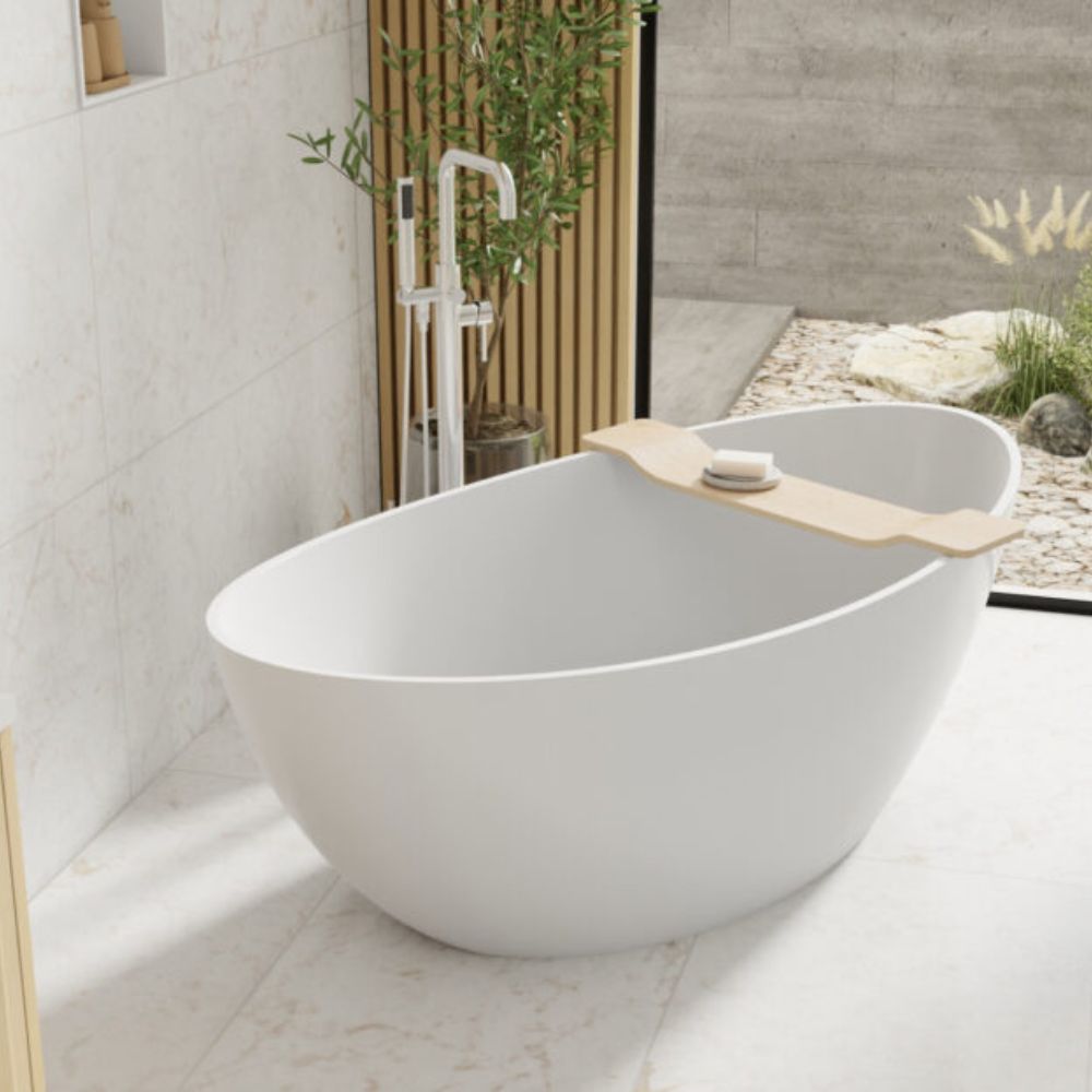 A freestanding oval bathtub in white with a light wood caddy holding a soap dish and bar of soap, set in a bright bathroom with tiled walls and a modern faucet.