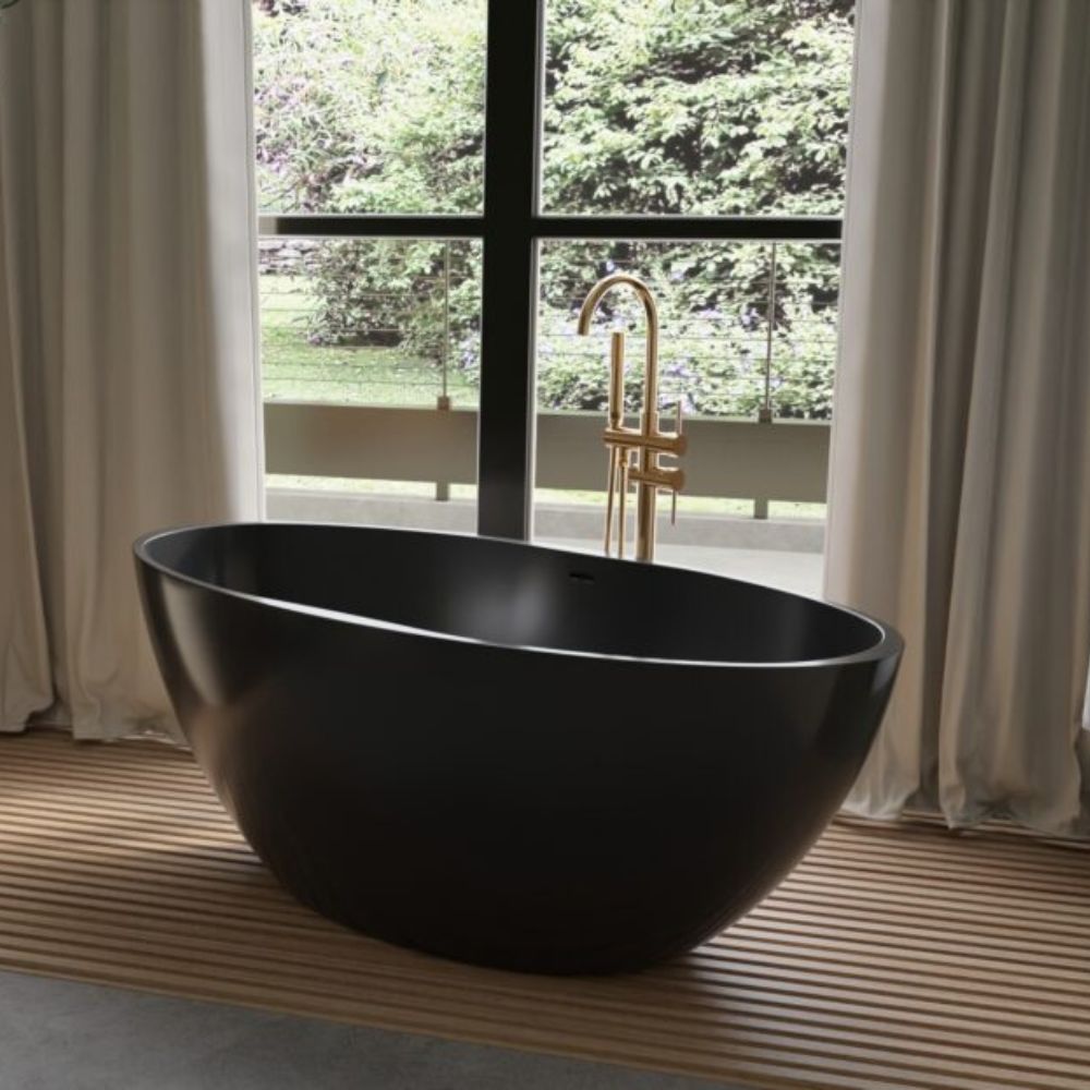 A matte black freestanding oval bathtub placed on a slatted wooden platform in front of a large window with beige curtains, with a gold faucet and an outdoor garden view.