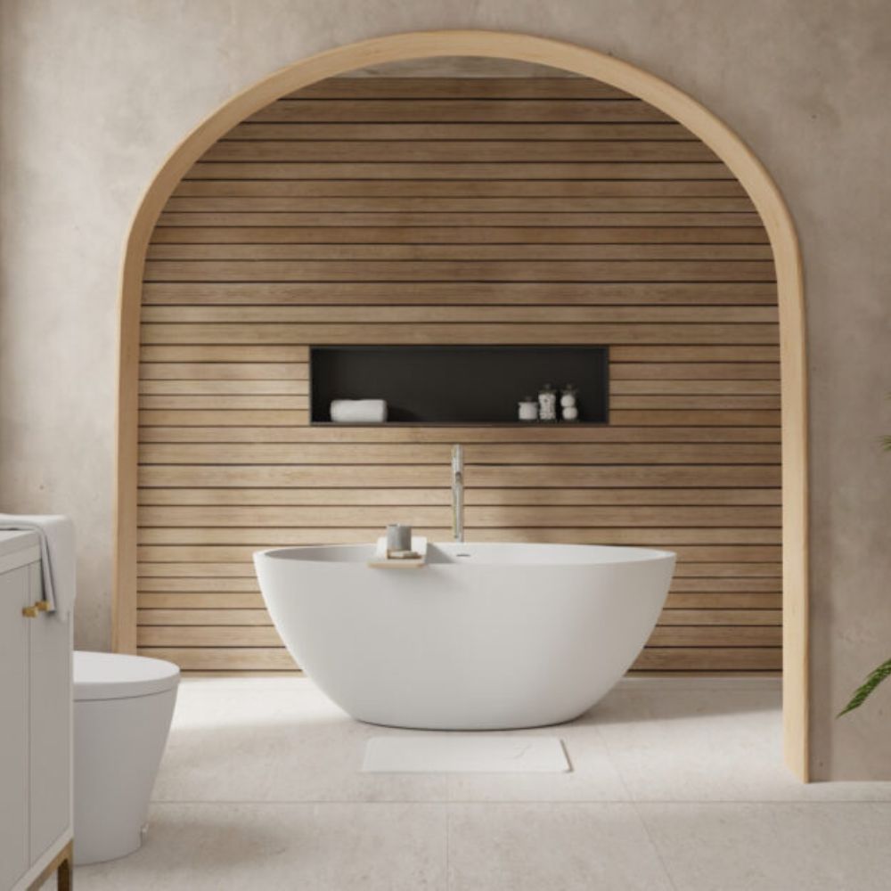 A freestanding oval white bathtub with a chrome faucet, set in a modern bathroom with a light wood slatted arched wall and textured beige plaster surroundings.