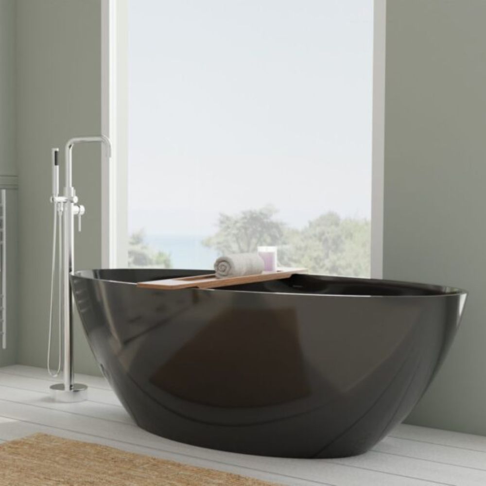 A modern freestanding oval bathtub in glossy black, shown in a bright bathroom setting with a tall window, a chrome floor-mounted faucet, and a small wooden tray holding a rolled towel.