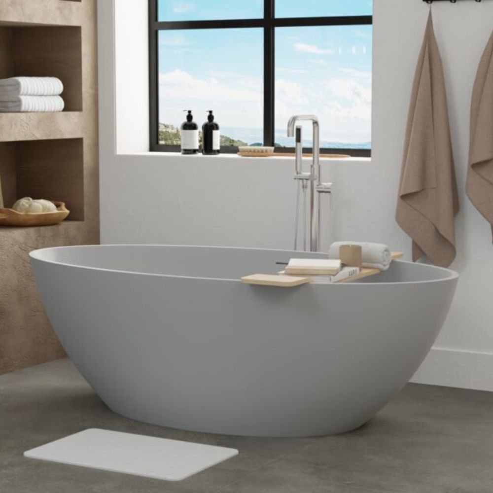 A modern freestanding oval bathtub in matte light gray, shown in a bright bathroom setting with a chrome faucet, bath mat, towels, and a window view.