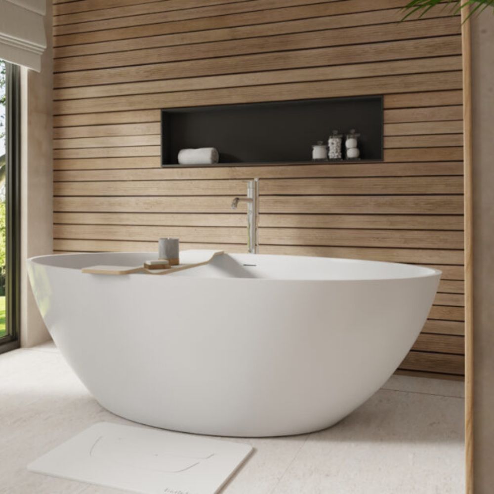 A modern freestanding oval bathtub in matte white, set in a minimalist bathroom with a light wood slat wall, a recessed shelf with toiletries, and a chrome faucet.