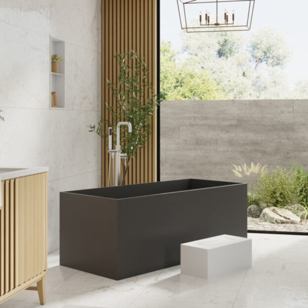 A modern freestanding rectangular bathtub in matte black, shown in a minimalist bathroom setting with a light wood vanity, a white stool, and a large window looking out to greenery.