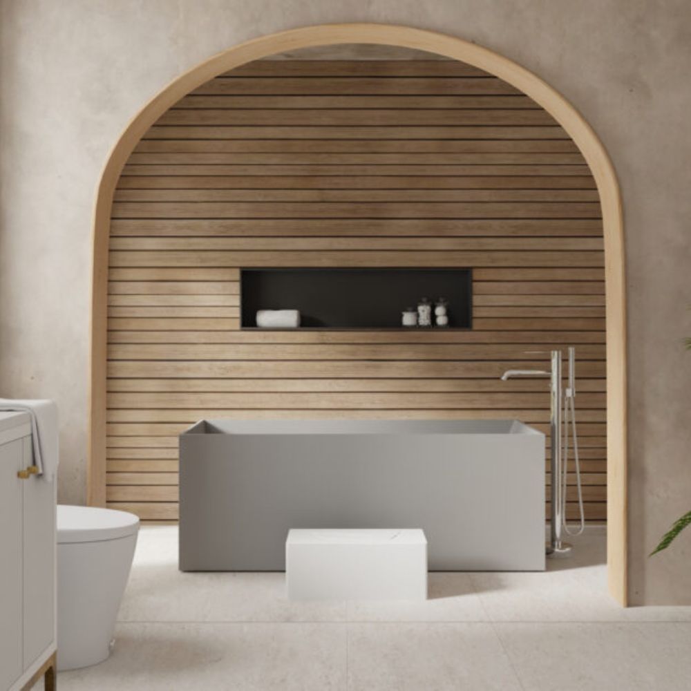 A modern freestanding bathtub in matte gray positioned in front of a warm wood slat wall with an arched frame, set in a minimalist bathroom with a white toilet and vanity visible.