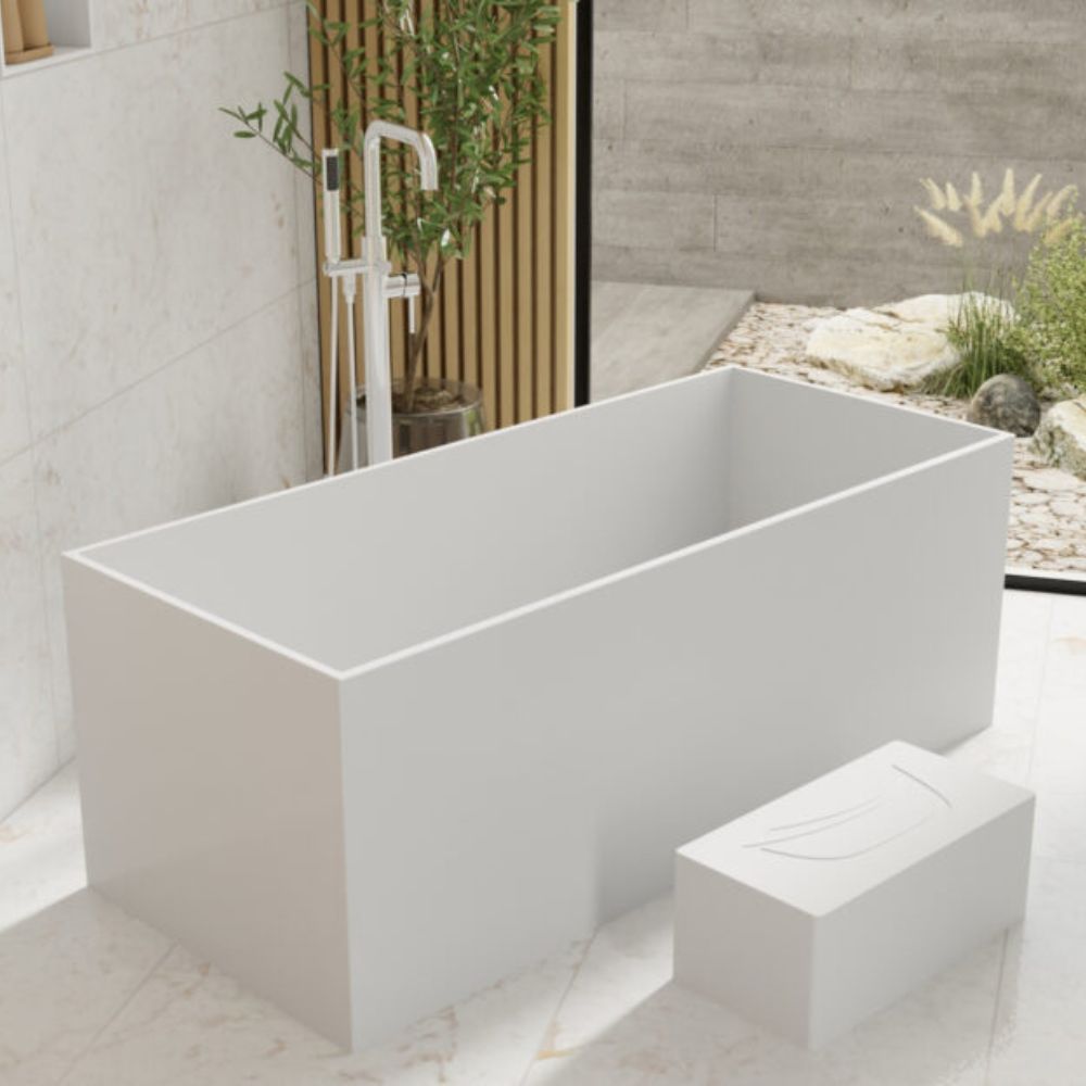 A modern freestanding rectangular bathtub in matte white, shown in a minimalist bathroom setting with a matching white footrest and a small outdoor garden area visible through a glass partition.