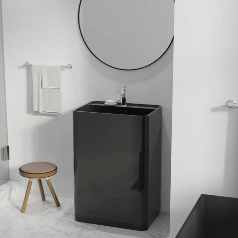 A modern glossy black rectangular pedestal sink with a chrome faucet, set in a minimalist bathroom with a round mirror, white towel, and a small wooden stool.