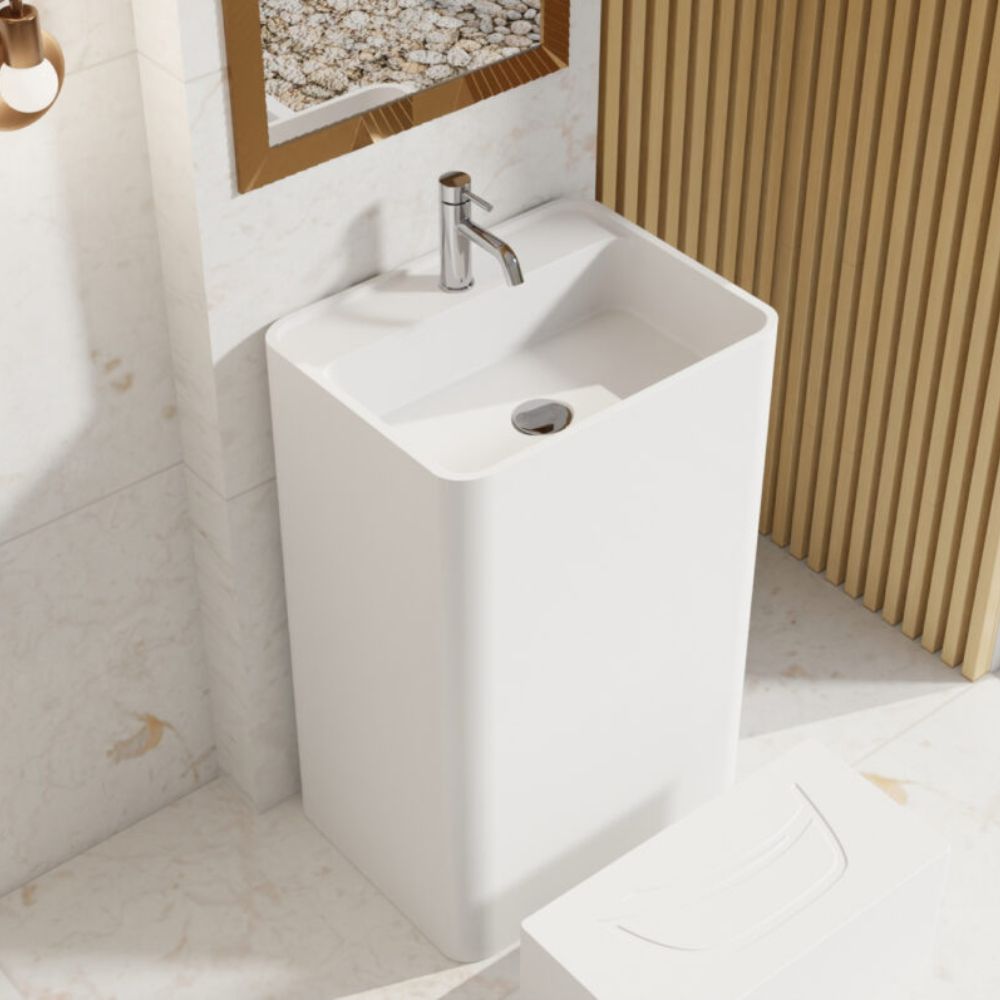A modern white pedestal bathroom sink with a chrome faucet, shown in a styled bathroom setting with marble tiles, a slatted wood wall panel, and a mirror.