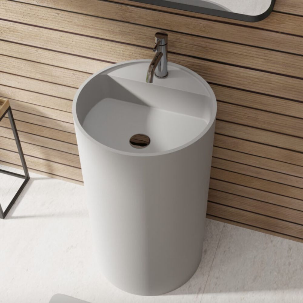 A modern cylindrical white pedestal sink with a chrome faucet, set against a light wood slat wall and white tiled floor.