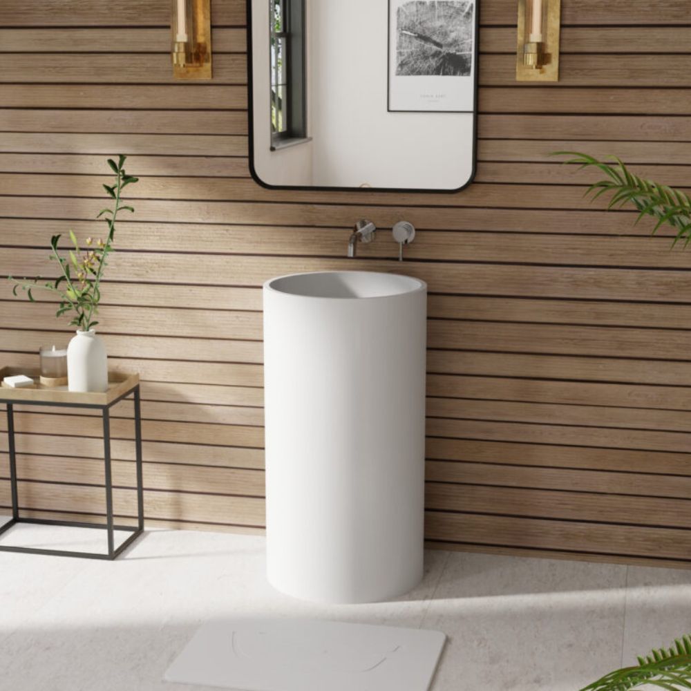 A modern white cylindrical pedestal sink in a bathroom setting with a wood slat wall, wall mirror, brass sconces, and a small side table.