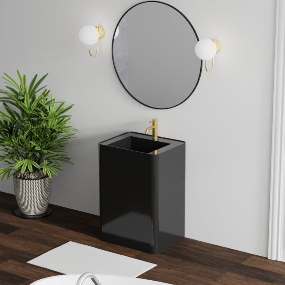 A modern black rectangular pedestal bathroom sink with a gold faucet, set against a light gray wall with a round mirror, two wall sconces, a potted plant, and a dark wood floor.