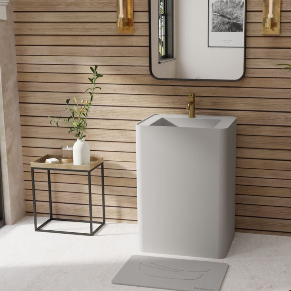 A modern gray pedestal sink with a matte finish and a gold faucet, set in a bathroom with a wood slat wall, a small side table with a vase, and a gray floor mat.
