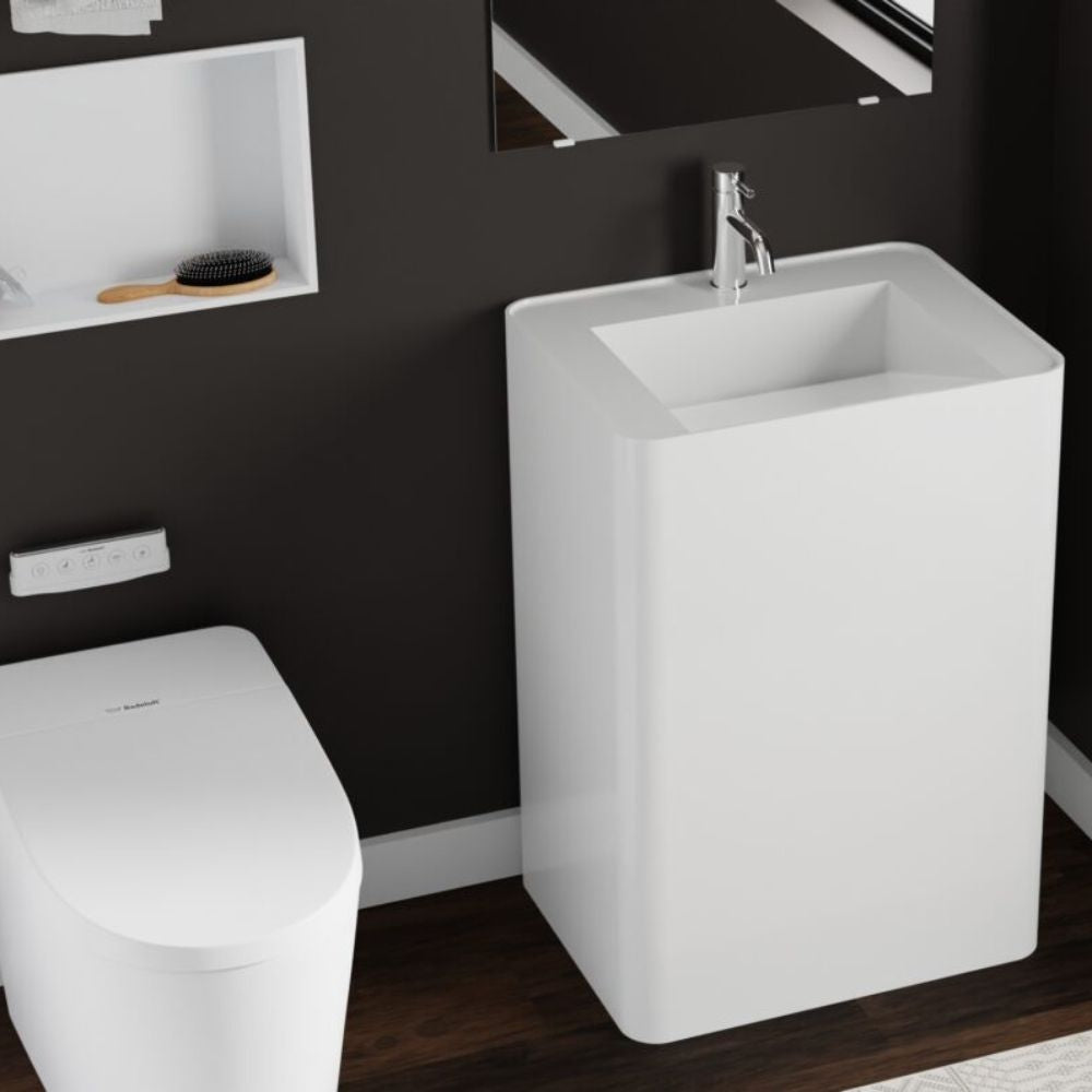 A modern white pedestal sink with a chrome faucet, shown in a bathroom setting next to a white toilet and a dark wall.