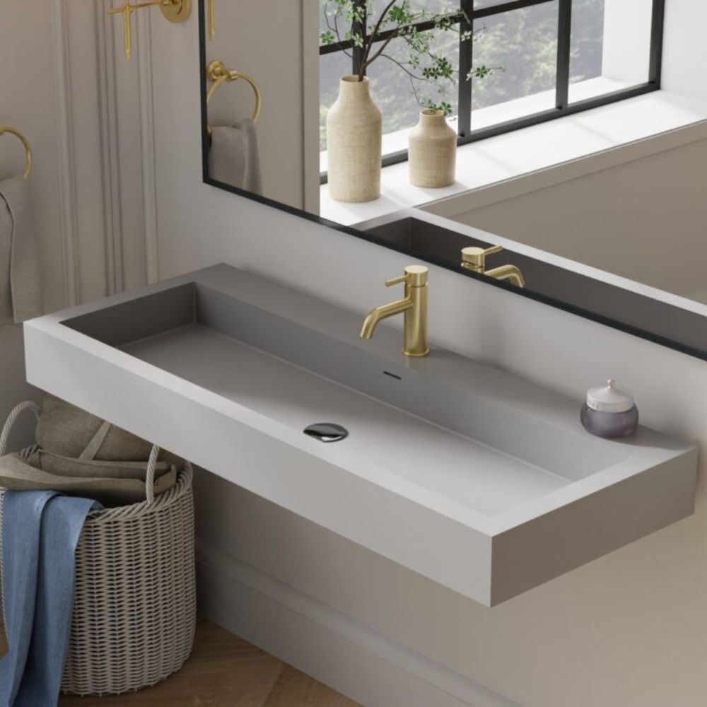 A modern wall-mounted rectangular sink in a light gray finish with a matte gray interior, paired with a brushed gold faucet. The sink is shown in a styled bathroom setting with a large mirror, gold towel rings, a wicker basket with towels, and decorative vases on the windowsill.